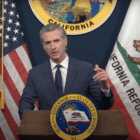 Govenor Gavin Newsom stands behind a podium, gesturing with his left hand. The California state flag, state seal, and an American flag hang behind him